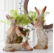 Three decorative rabbits with bows in a home setting with plants.