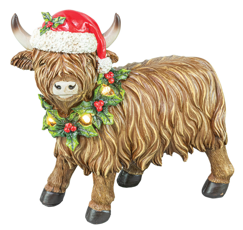 A decorative Highland cow figurine with long shaggy brown fur, black hooves, a red Santa hat with white trim, and a green holly wreath with red berries and warm glowing lights, shown on a plain white background.