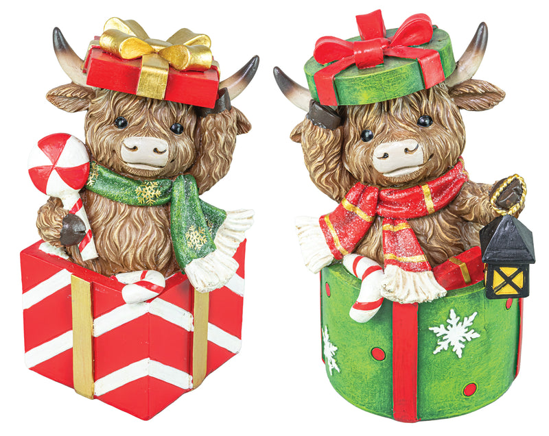 Two Highland cow holiday figurines sitting in gift boxes, decorated with red and green wrapping, scarves, candy canes, and a small lantern, shown on a white background.