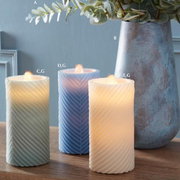 Three wavy-textured candles on a table with a vase of flowers in the background.