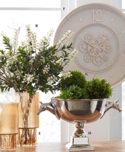 Decorative silver urn with greenery, vase with flowers, and clock on a table.