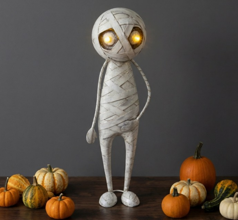 Mummy figurine with glowing eyes surrounded by pumpkins on a wooden surface.