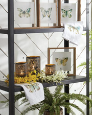 A styled interior shelf display featuring butterfly-themed framed prints, white towels with butterfly illustrations, and decorative metal candle holders with tree silhouettes and yellow flowers against a white paneled wall.