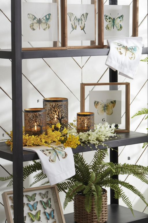 A styled interior shelf display featuring butterfly-themed framed prints, white towels with butterfly illustrations, and decorative metal candle holders with tree silhouettes and yellow flowers against a white paneled wall.