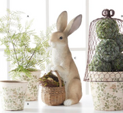 Decorative scene with a plush rabbit, basket, and potted plants on a white surface.