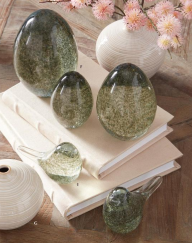Set of green glass eggs on a wooden surface with a vase and flowers in the background