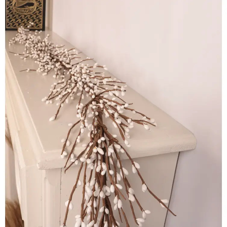 A decorative faux willow branch garland with white fluffy buds draped over a white fireplace mantel against a light wall.