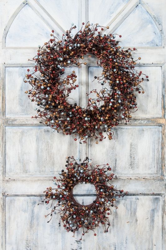 Two rustic berry wreaths with red and silver berries hanging on a distressed white wooden door.