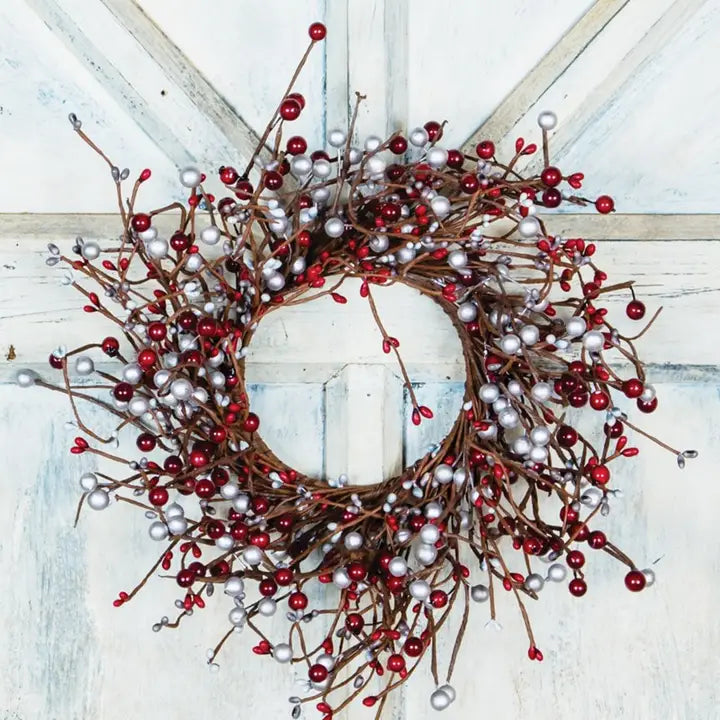 A festive holiday wreath with brown branches, red berries, and silver ornaments hanging on a distressed white wooden door.