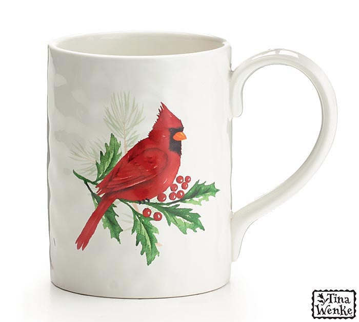 A white ceramic mug featuring a painted illustration of a red cardinal perched on a holly branch with green leaves and red berries, shown on a white background.