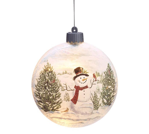 A round glass Christmas ornament featuring a painted winter scene with a snowman, evergreen trees, and a small bird, shown hanging against a white background.