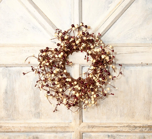 A rustic berry wreath with dark red berries and cream-colored pods on a brown vine base, hanging on a distressed white paneled wooden door.