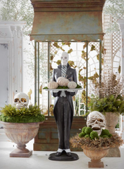 Halloween-themed decoration with skeletons, pumpkins, and decorative urns in a garden setting.