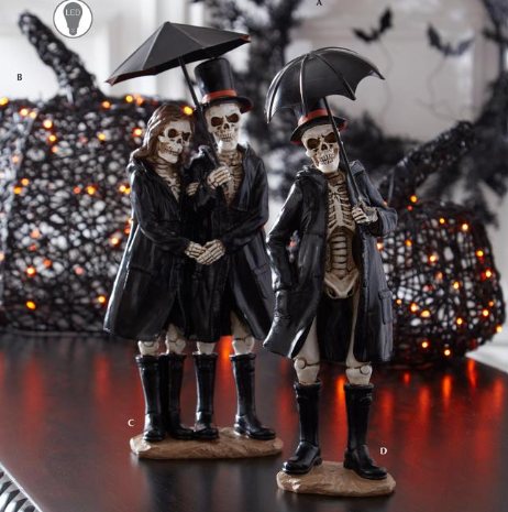 Two Halloween skeleton figurines dressed in black Victorian-style coats and top hats, each holding a black umbrella, displayed on a dark reflective surface with spooky decorative lights in the background.