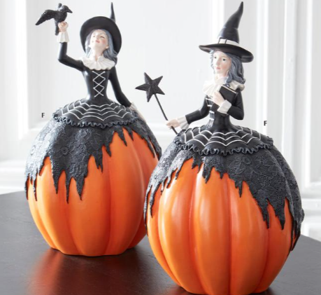 Two Halloween pumpkin decorations with witch figurines, featuring one holding a bird and the other holding a star wand, displayed on a dark tabletop against a bright indoor background.