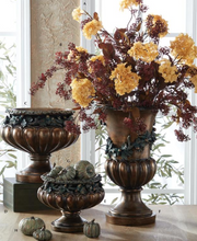Three bronze-finished decorative urns arranged on a table in a bright room, with the largest urn holding a floral arrangement of yellow and burgundy hydrangea-like blooms. Small pumpkins and green gourds sit in the smaller urns, and a window and textured wall are visible in the background.