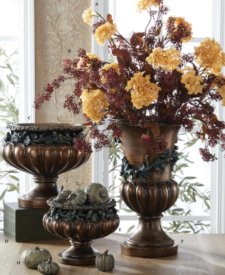 Three bronze-finished decorative urns arranged on a table in a bright room, with the largest urn holding a floral arrangement of yellow and burgundy hydrangea-like blooms. Small pumpkins and green gourds sit in the smaller urns, and a window and textured wall are visible in the background.