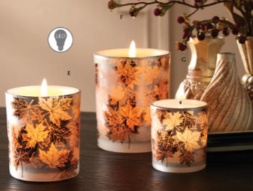 Three lit taper candles with a warm orange and gold leaf pattern, displayed on a dark wooden surface with a decorative ceramic vase in the background.