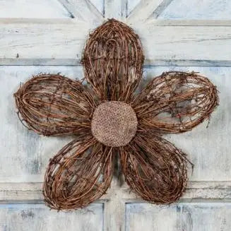 A rustic five-petal flower decoration made from brown woven vine branches, with a burlap-covered center, displayed against a distressed white wooden background.