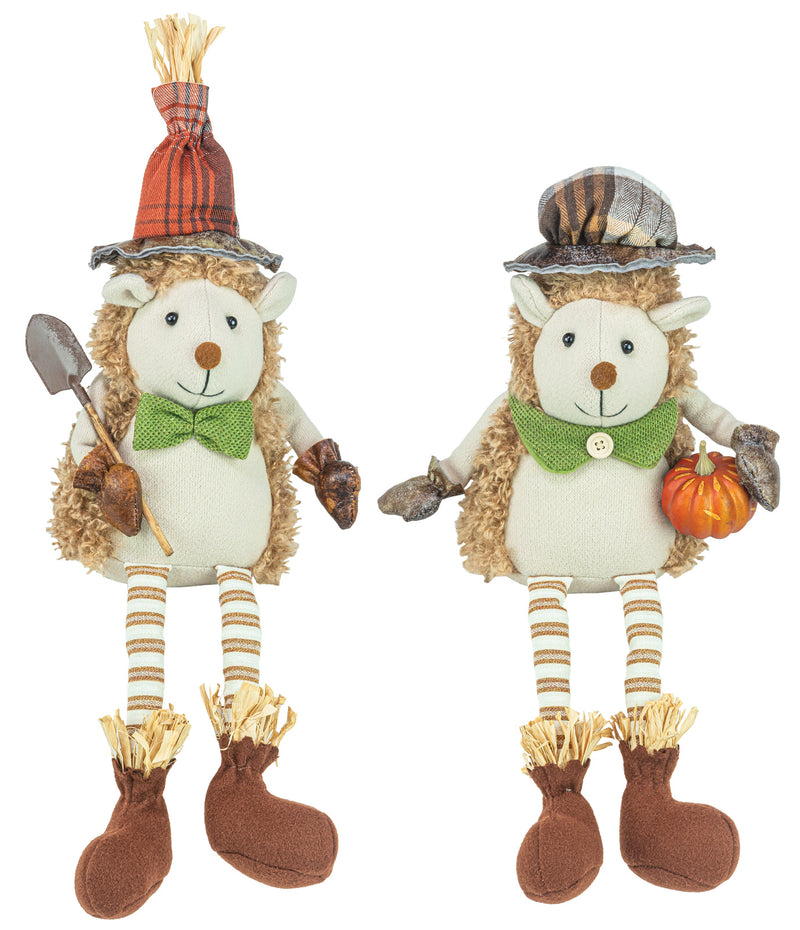 Two autumn-themed hedgehog plush toys standing side by side on a white background. One wears an orange plaid hat with a straw top, a green bow tie, striped legs, and brown boots while holding a small shovel.