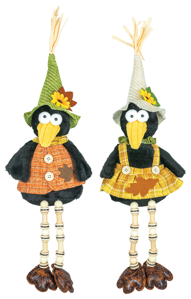 Two decorative crow-shaped hanging ornaments with long legs, wearing autumn-themed outfits. One has a green hat and orange plaid vest, and the other has a white hat and yellow plaid dress, both on a plain white background.