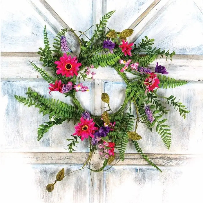 A decorative floral wreath with green ferns and bright pink, purple, and red flowers, displayed against a distressed white wooden door.