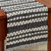 A woven table runner with a black and cream striped pattern draped over a wooden table, showing its textured, braided weave detail.