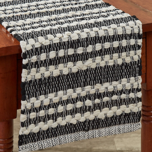 A woven table runner with a black and cream striped pattern draped over a wooden table, showing its textured, braided weave detail.
