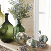 Decorative glass vases and mirrored ornaments arranged on a windowsill with dried flowers and pinecones. The set includes a large dark green glass bottle, a tall green glass vase with floral stems, and three round mirrored ornaments with a green tree pattern.