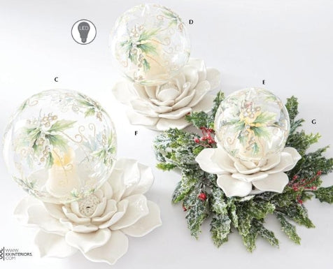 Three clear glass snow globes with painted winter scenes, displayed on white flower-shaped bases. One larger version is on a white flower base, and two smaller versions are arranged with green frosted holly and red berries.