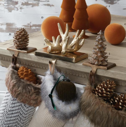 A styled holiday tabletop scene with orange decorative spheres, pinecones, a gold antler centerpiece, and knit stockings hanging from brass hooks. No person or animal is present, and the setting reads as a styled product display rather than a real lived-in environment, so Product/Packaging fits better than Lifestyle.