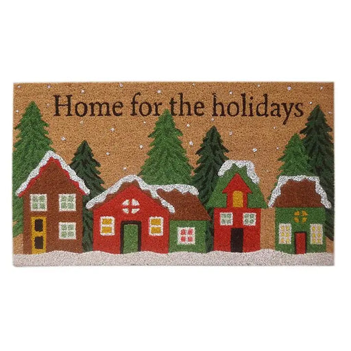 A rectangular coir doormat with a festive winter village design. The natural brown coir background features the text 'Home for the holidays' in black, decorated with red, green, and brown houses, white snow, and green pine trees.