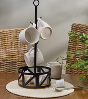 Black metal mug tree with white mugs on a wooden table with a wicker chair and plant in the background.