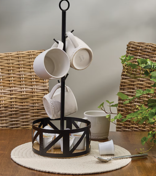 Black metal mug tree with white mugs on a wooden table with a wicker chair and plant in the background.
