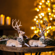 Two deer figurines with wreaths on a wooden table in front of blurred lights