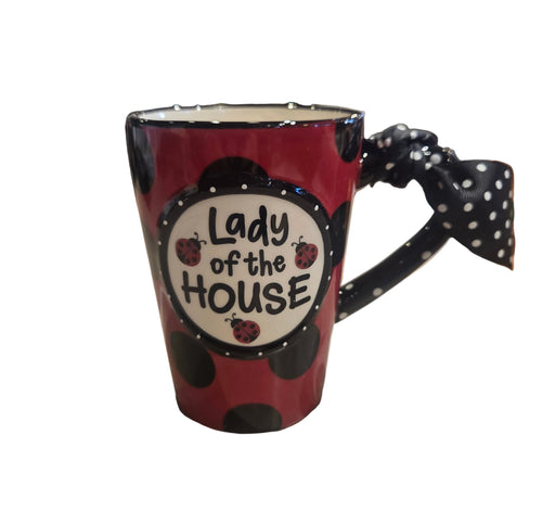 A red ceramic mug with black polka dots and a black-and-white polka-dot bow handle, featuring a white oval label that reads 'Lady of the HOUSE' with ladybug illustrations. The mug is photographed on a stone countertop in a store setting with blurred background shelves.