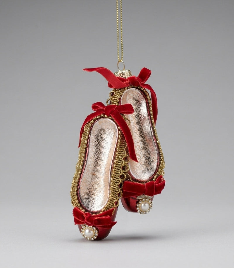A hanging ornament shaped like a pair of red velvet ballet flats with gold trim, metallic pink interiors, red ribbon bows, and pearl accents, shown against a neutral gray background.