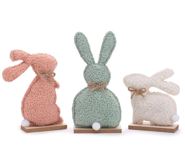 Three textured fabric bunny figurines in pink, sage green, and cream, each tied with a small jute bow and standing on wooden bases against a white background.
