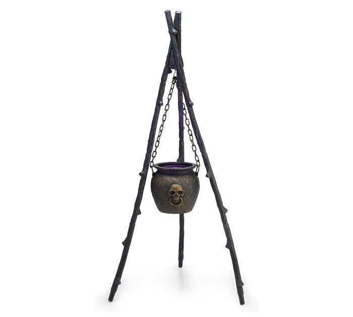 Halloween decoration featuring a black metal cauldron with a skull detail hanging from chains on a tripod stand made of black branches.