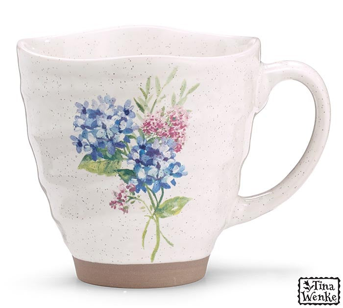 A speckled off-white ceramic mug with a watercolor floral print of blue and pink flowers with green leaves, featuring a brown glazed base and a C-shaped handle. A 'Tina Wenke' brand logo is visible in the bottom right corner.