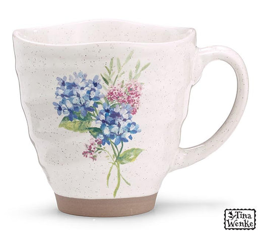 A speckled off-white ceramic mug with a watercolor floral print of blue and pink flowers with green leaves, featuring a brown glazed base and a C-shaped handle. A 'Tina Wenke' brand logo is visible in the bottom right corner.
