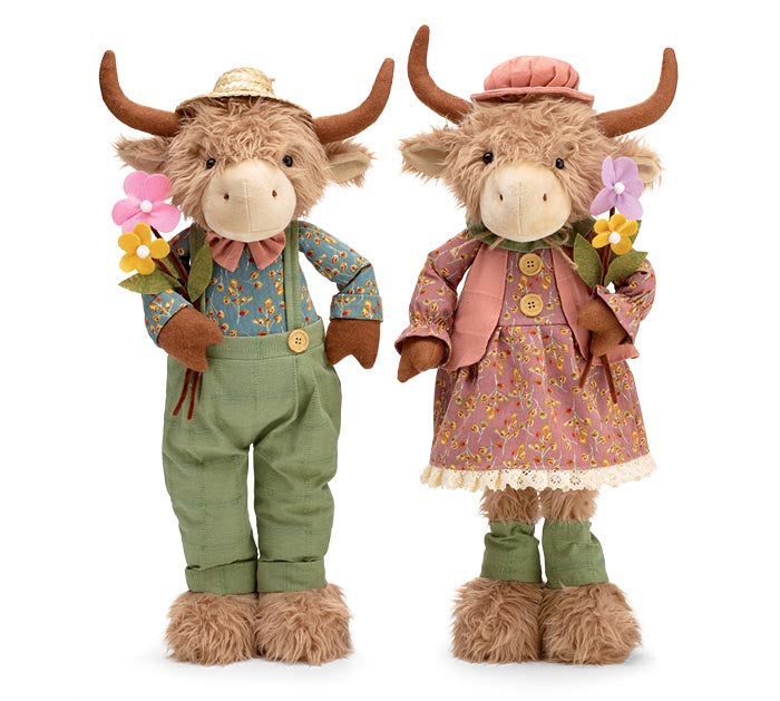 Two plush Highland cow dolls standing side by side on a white background. One doll wears green overalls with a blue floral shirt and a straw hat, holding pink and yellow flowers.