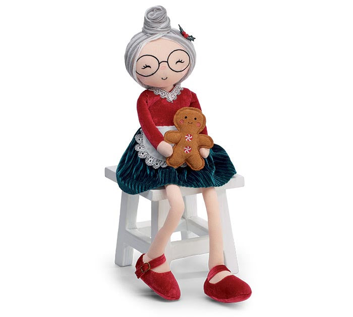 A festive holiday figurine of an elderly woman with gray hair and glasses, wearing a red top, dark green skirt, and red shoes, sitting on a white stool and holding a gingerbread man.