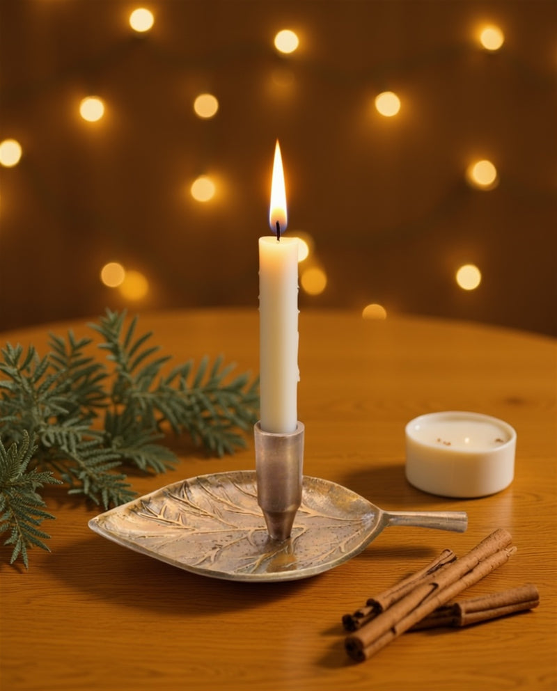 Candle in a leaf-shaped holder with cinnamon sticks and a small candle on a wooden surface, blurred lights in the background.