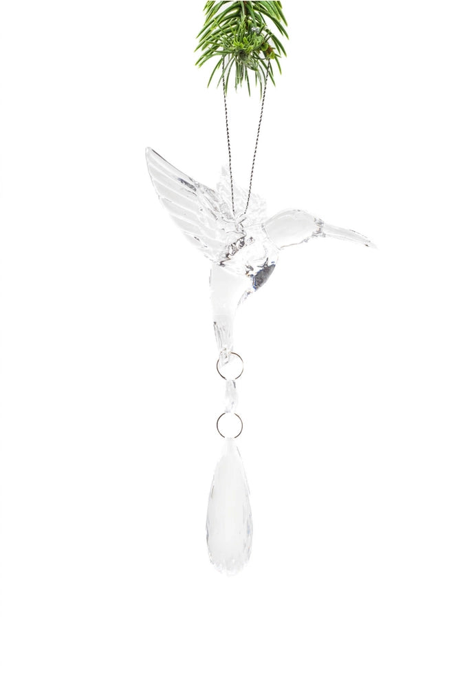 A clear acrylic or glass hummingbird Christmas ornament hanging from a silver chain, with a green pine branch at the top and a teardrop-shaped clear drop below, shown against a white background.