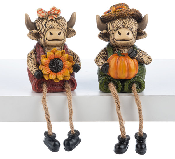 Two decorative cow figurines with autumn-themed accessories on a white shelf.