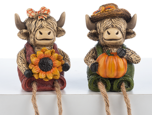 Two decorative cow figurines with autumn-themed accessories on a white shelf.