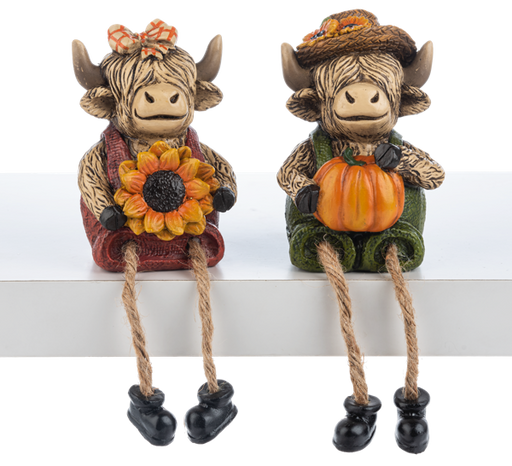 Two decorative cow figurines with autumn-themed accessories on a white shelf.