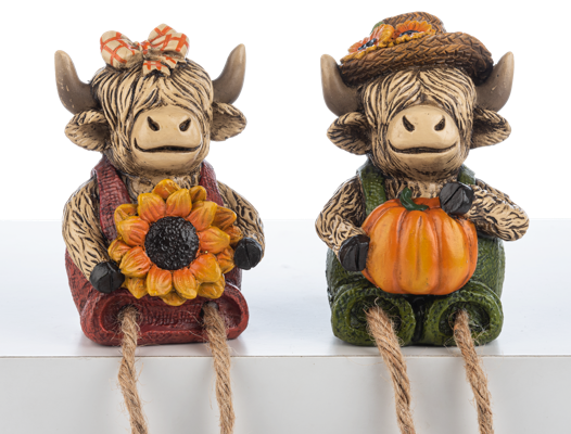 Two decorative cow figurines with autumn-themed accessories on a white shelf.