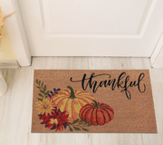 Doormat with pumpkins and 'thankful' text on a wooden floor.